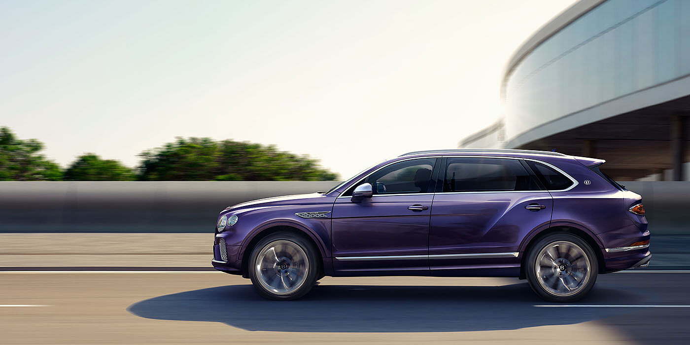 Bentley Leicester Bentley Bentayga Extended Wheelbase Mulliner luxury SUV profile in Tanzanite Purple paint, driving on an country road past trees.