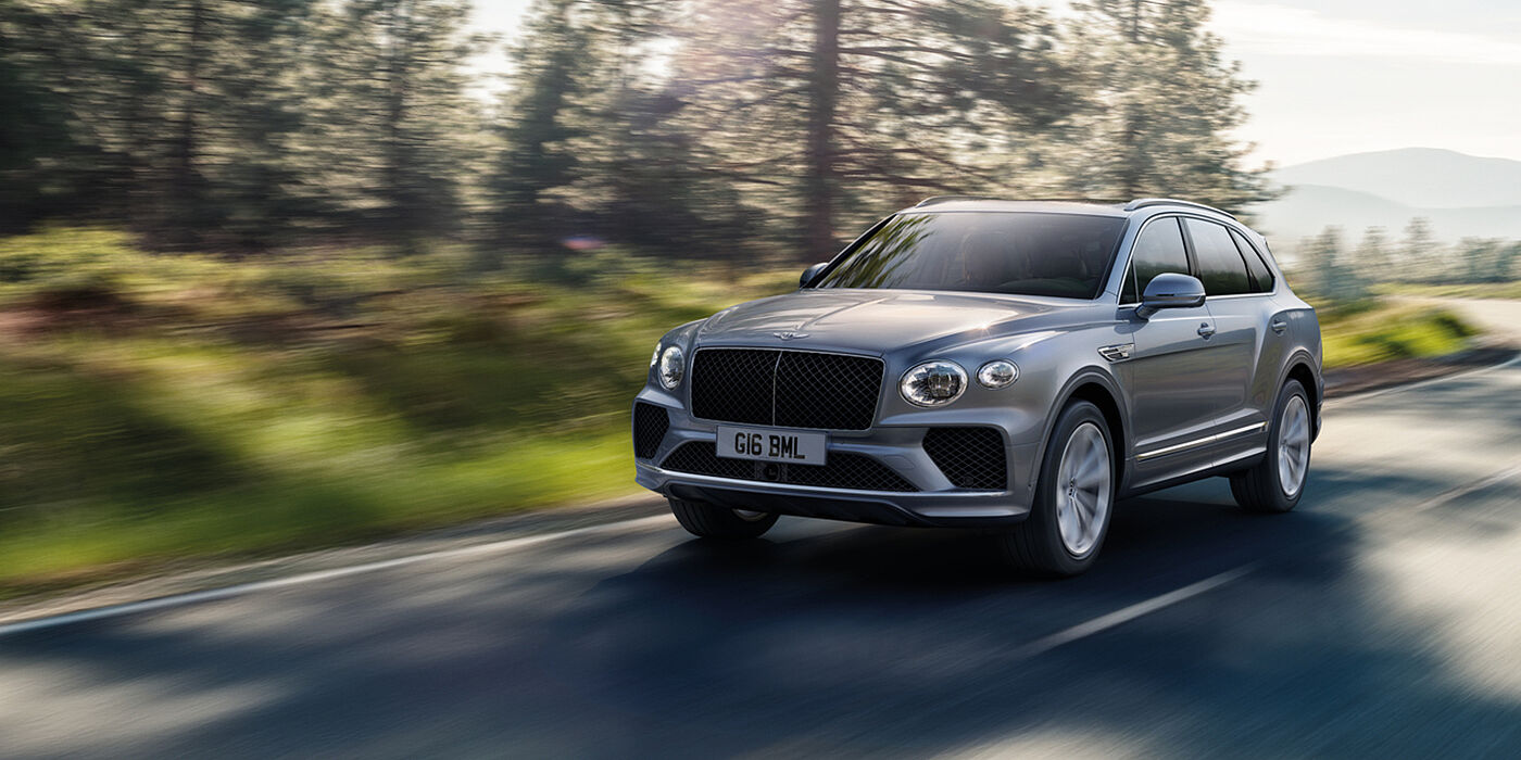Bentley Leicester Bentley Bentayga luxury SUV front three quarter in Moonbeam paint, driving on an country road past trees.