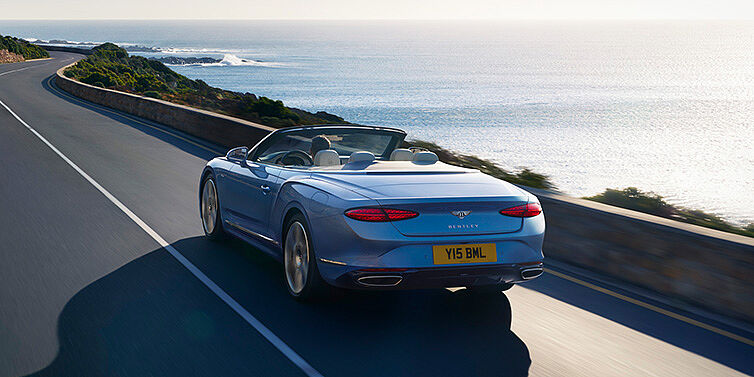 Bentley Leicester New Bentley Continental GTC Azure convertible rear view in Light Windsor blue paint with 22 inch ten spoke wheels driving dynamically on an ocean road in the sunshine