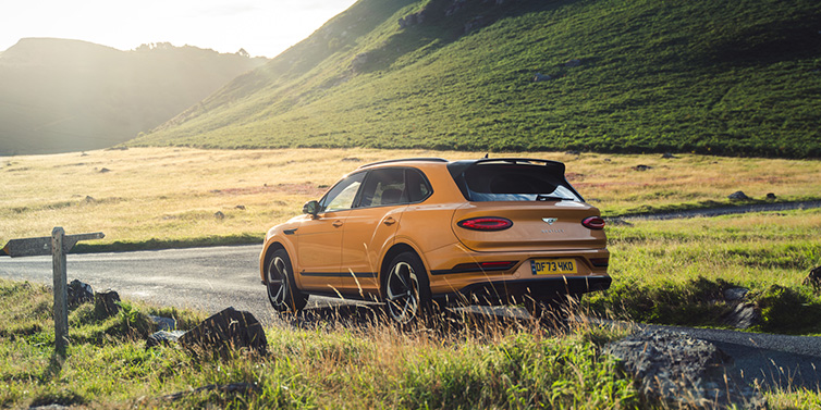 Bentley Leicester Bentley Bentayga S SUV rear three quarter in Sunburst Gold paint on a road surrounded by green hills