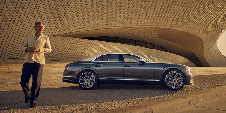 Bentley Leicester Bentley Flying Spur Mulliner sedan in Rose Gold over Magnetic duotone paint parked by a curved wall with person in suit jacket standing on the left